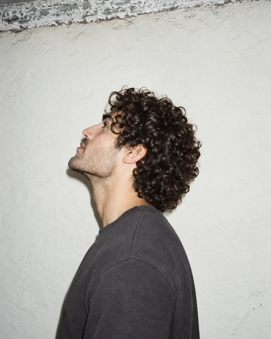 Side profile portrait with dense dark curls against a pale textured wall